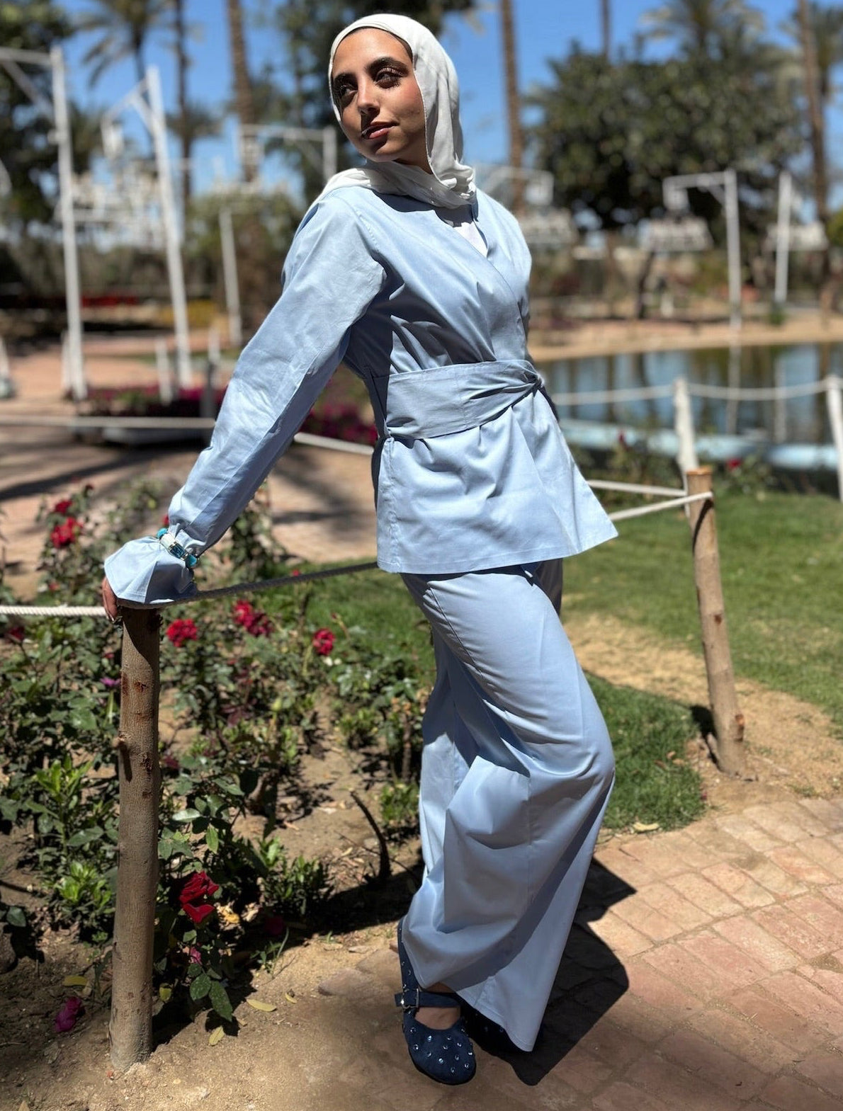 Woman in a light blue outfit standing outdoors with greenery and a pond in the background